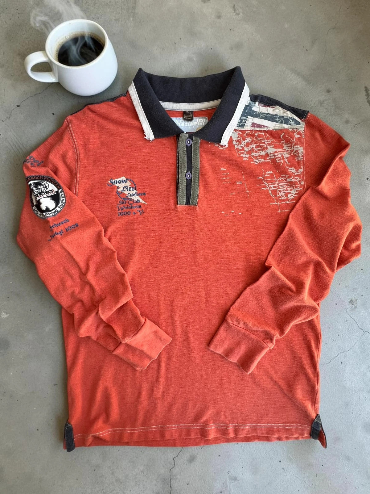 Napapijri orange long sleeve polo with embroidered patches and "Forever Wild and Free" graphic.