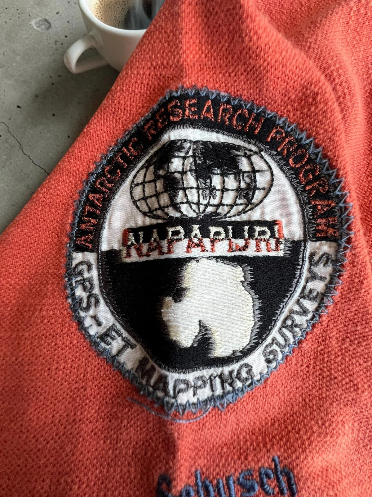 Napapijri orange long sleeve polo with embroidered patches and "Forever Wild and Free" graphic.